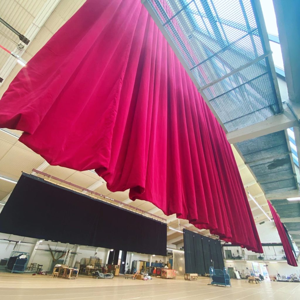 Großer roter Bühnenvorhang hängt in einer geräumigen Halle, im Hintergrund weitere schwarze Vorhänge und Arbeitsmaterial.