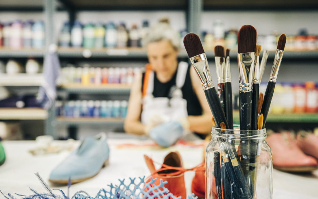 Brushes in a glass are in the foreground, while a blurred person works on a craft project in the background.