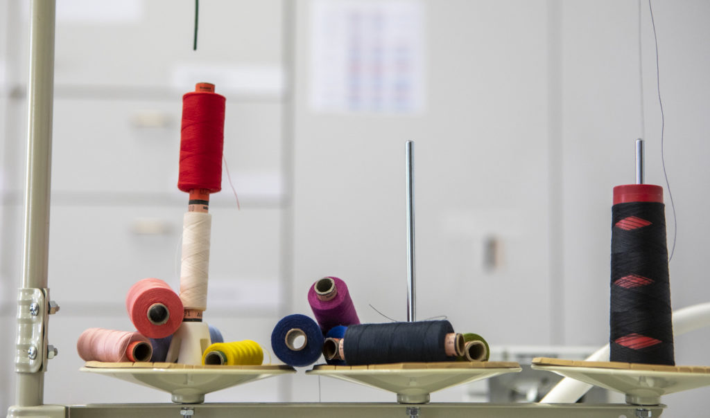 Various colourful spools of thread stand and lie next to each other on white holders in a workshop or sewing environment.