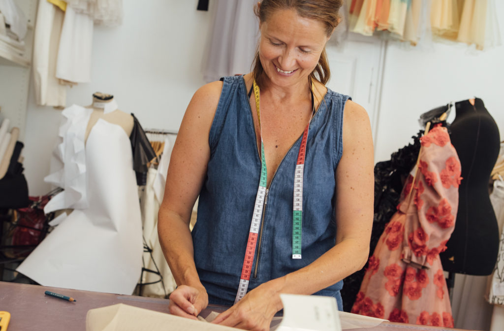 Eine lächelnde Frau in einem Schneideratelier arbeitet an Stoff, mit Maßband um den Hals und Kleidern im Hintergrund.