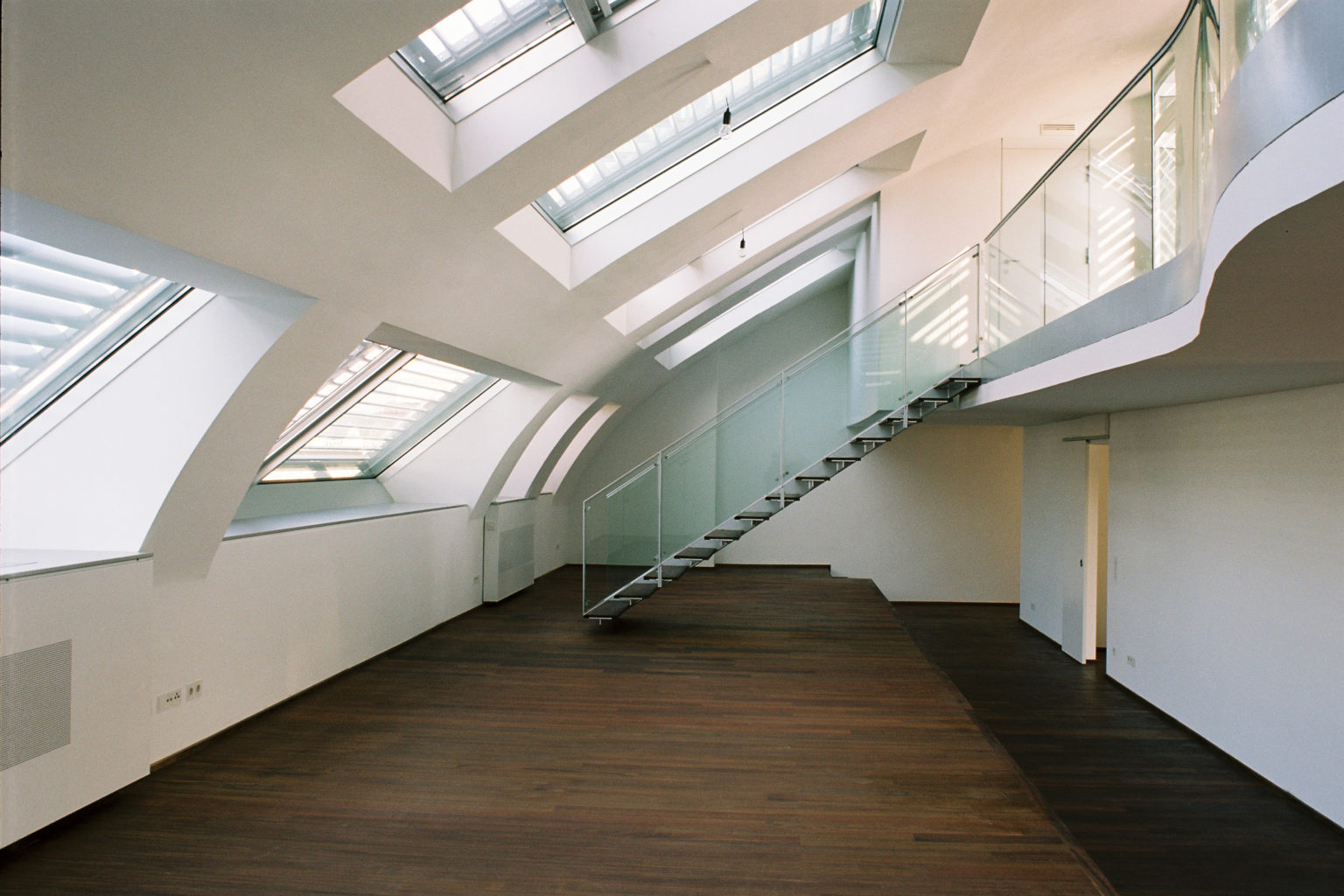 Large, modern attic room with wooden floor, arched windows and a staircase with glass banisters.