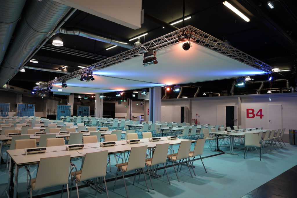 Ein großer, moderner Konferenzraum mit vielen weißen Stühlen und Tischen, Deckenbeleuchtung und dem Schild „B4“ an der Wand.