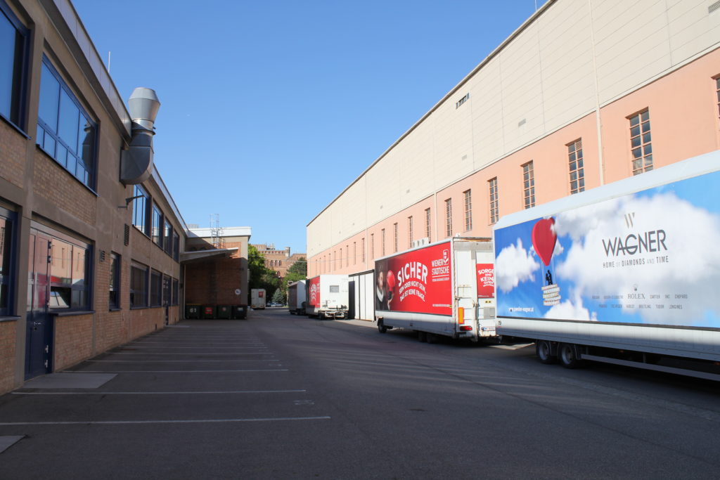 Lkw mit Werbeplakaten parken nebeneinander in einer sonnigen Industriehofeinfahrt zwischen Backstein- und Industriegebäuden.
