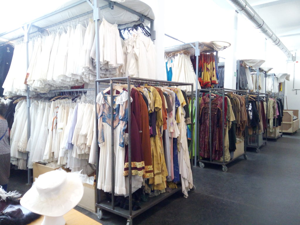 Mehrere Kleiderständer mit historischen Kostümen in verschiedenen Farben stehen in einem hellen, ordentlichen Raum.