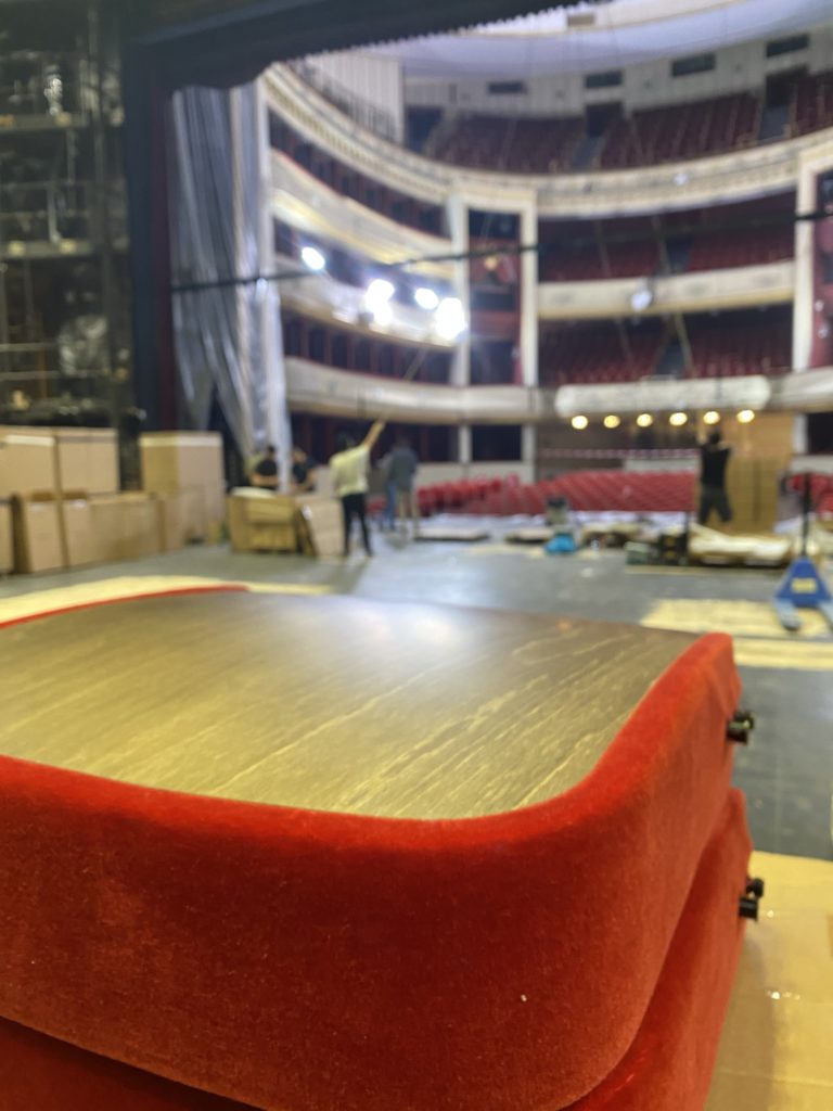 Red chair cushions in the foreground of a theatre stage with people and spotlights working on the stage in the background.