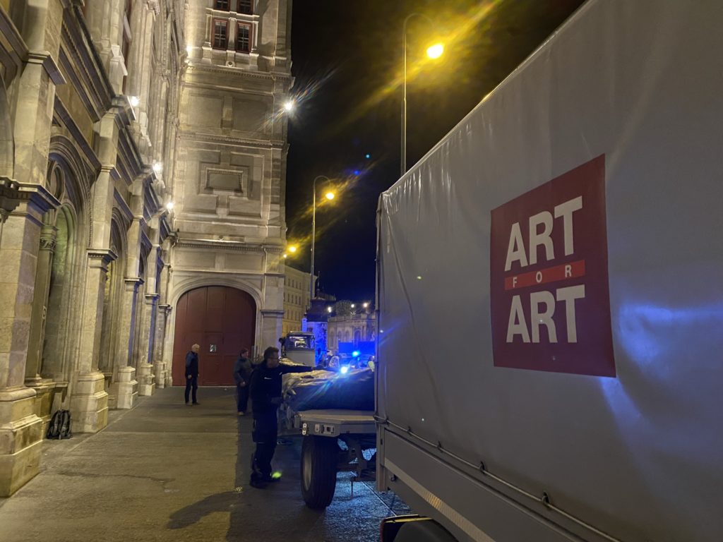 A lorry with the words „ART FOR ART“ is parked in front of a historic building at night, with several people standing next to it.