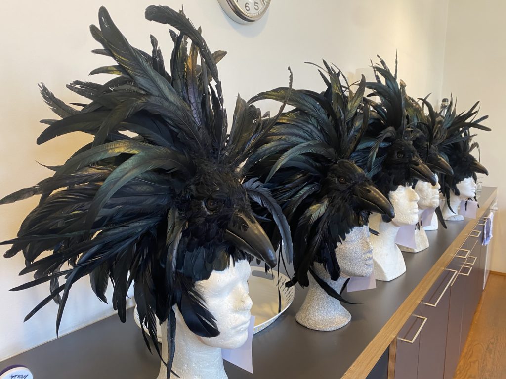 Mehrere Schaufensterpuppen tragen auffällige schwarze Kopfbedeckungen mit großen Federn, die wie Rabenköpfe geformt sind.