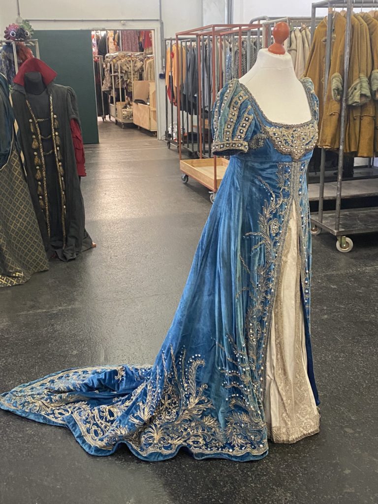 An elaborate, historic blue dress with silver embellishments and a long train on a dressmaker's dummy.