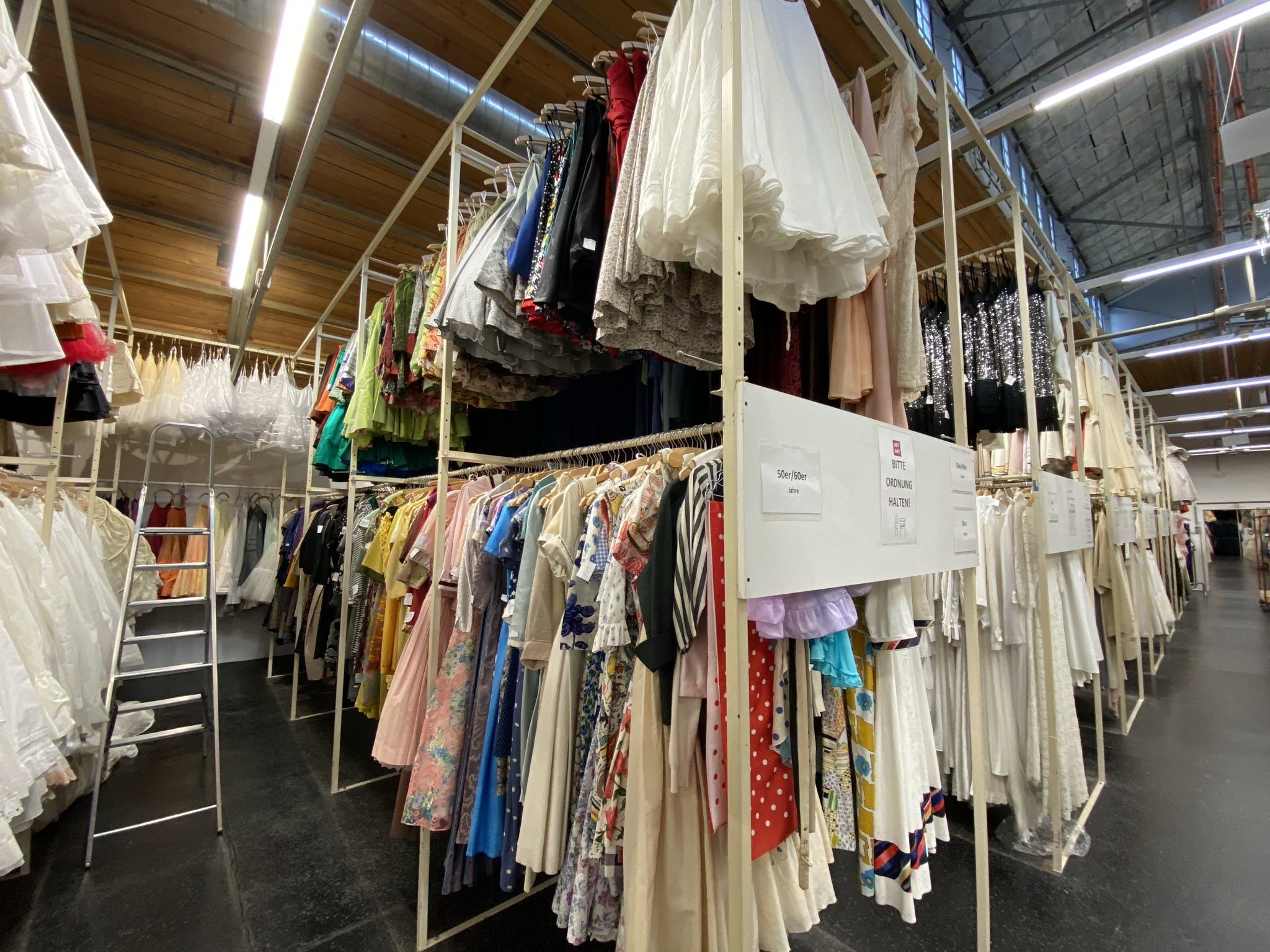 Große Gerüste mit zwei Kleiderstangen übereinander stehen in einer großen Halle. Auf ihnen hängen bunte Kleider  aus de 50er und 60er Jahren.
