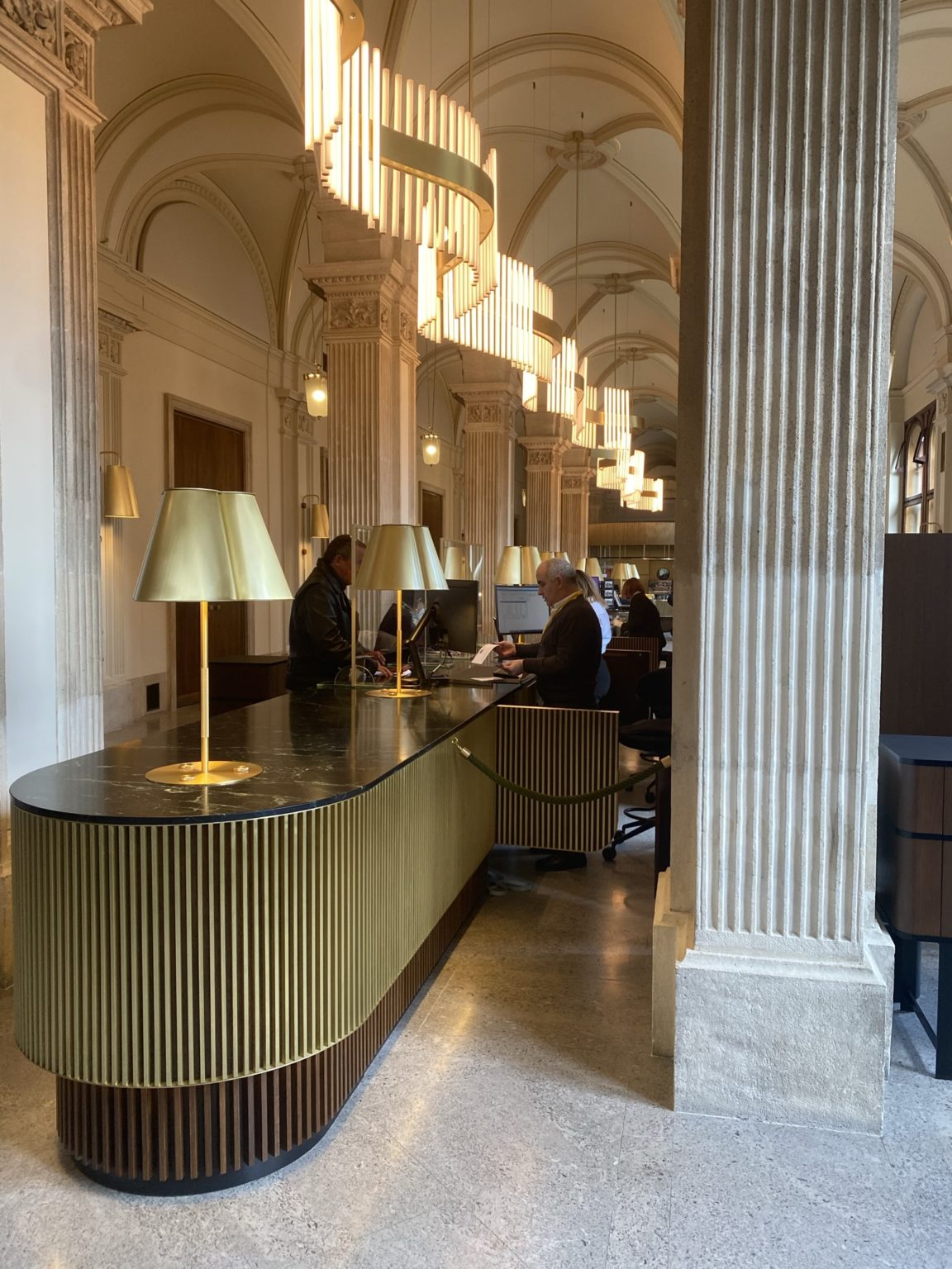 Mehrere Personen arbeiten an Schreibtischen mit goldenen Lampen in einer eleganten Halle mit hohen Decken und Säulen.