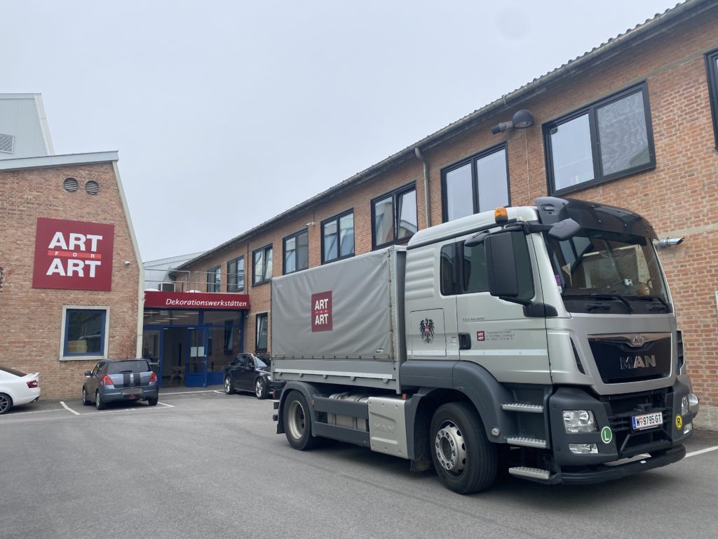 Ein LKW mit der Aufschrift „ART ART“ steht vor einem Backsteingebäude mit großem Schild und mehreren geparkten Autos.