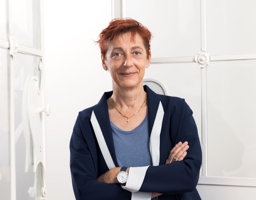 Frau mit kurzem, rötlichem Haar und blauem Blazer steht mit verschränkten Armen und lächelt leicht vor heller Wand.