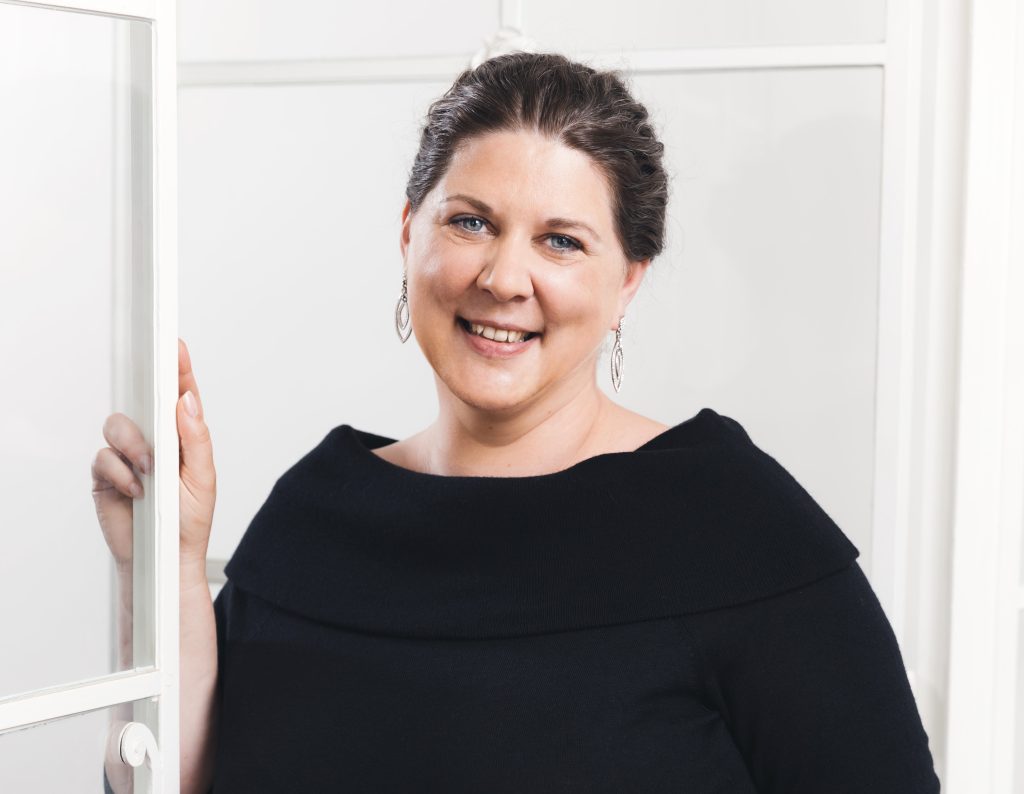 Frau mit dunklen Haaren und Ohrringen lächelt freundlich in eleganter schwarzer Kleidung vor einem hellen Hintergrund.