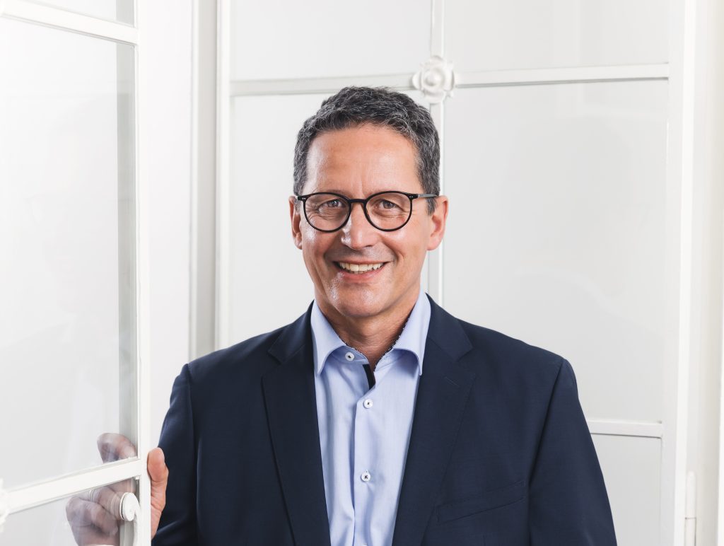 Ein lächelnder Mann mit Brille und dunklem Anzug steht vor einem Fenster mit weißem Rahmen.