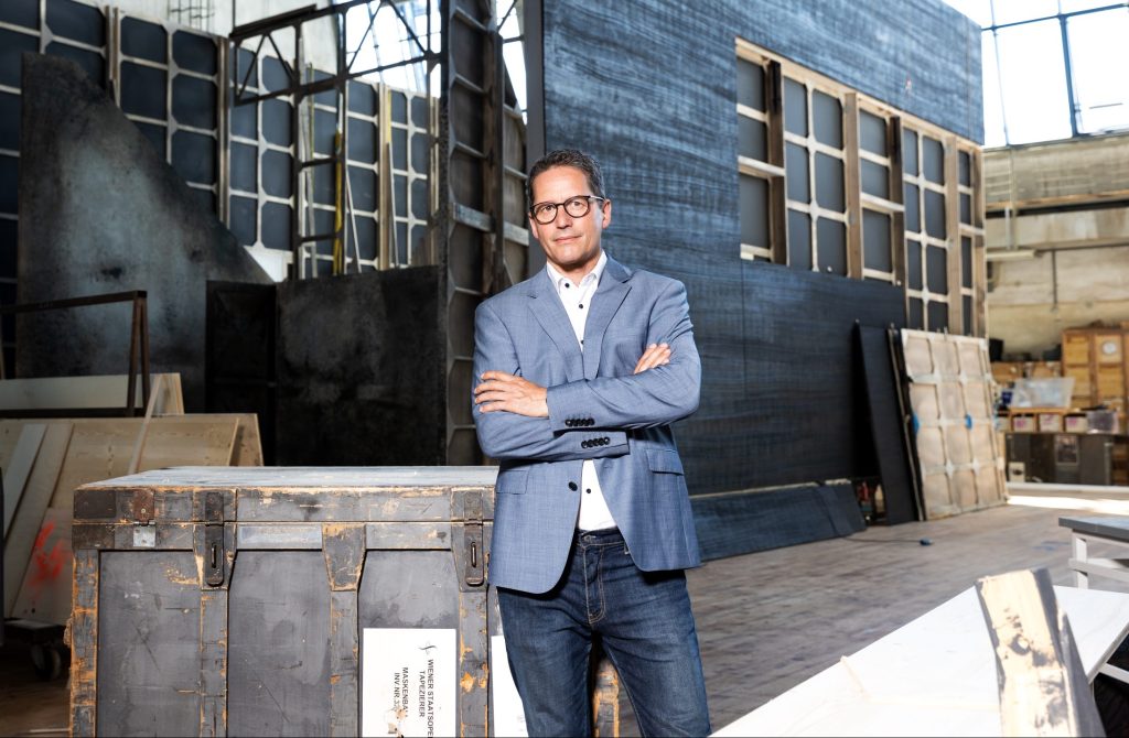 Ein Mann im blauen Sakko steht mit verschränkten Armen in einer Industriehalle vor einer großen Holzkiste und Bühnenkulissen.