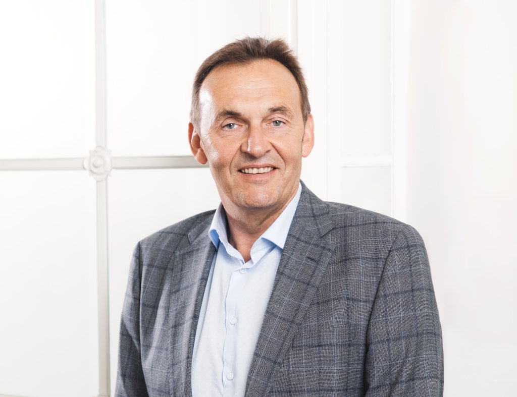 Ein lächelnder Mann in einem karierten Sakko und hellblauem Hemd steht vor einem hellen Hintergrund.