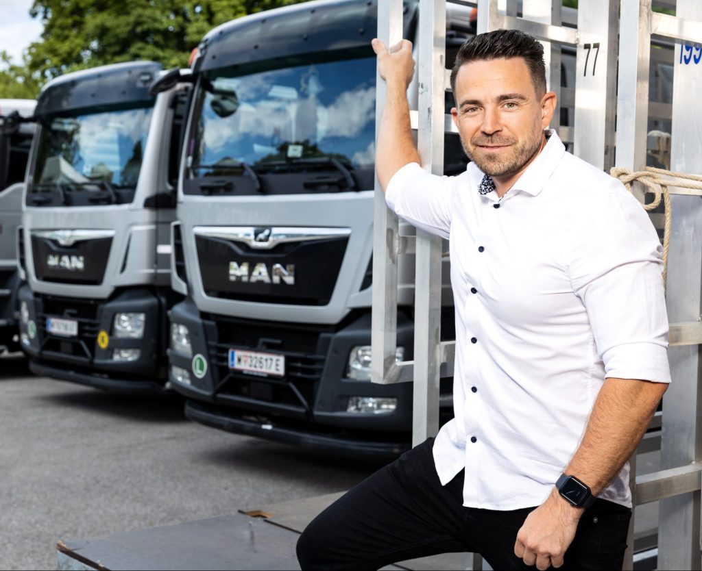 A man in a white shirt stands in front of several MAN lorries and smiles into the camera.