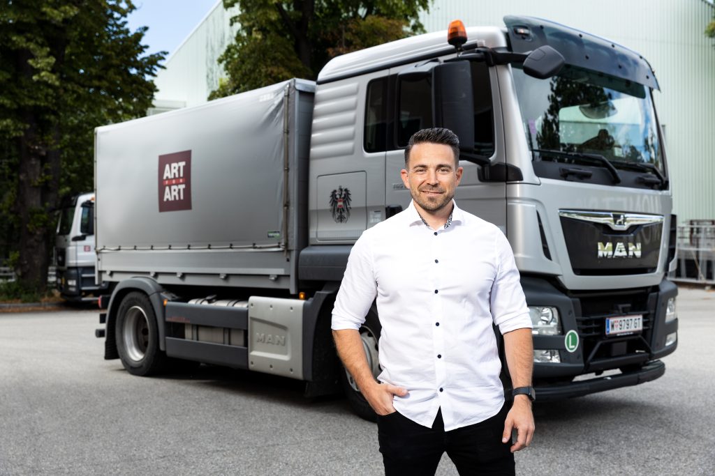 Ein Mann mit weißem Hemd steht lächelnd vor einem silbernen LKW mit der Aufschrift "ART ART" auf der Plane.