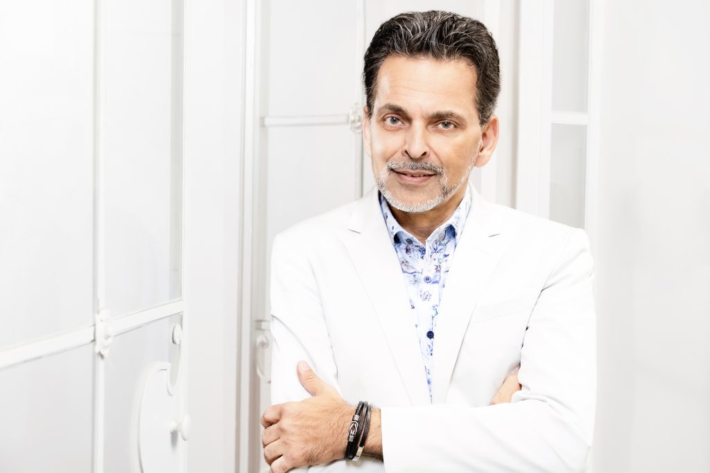 Ein Mann mit dunklen Haaren und Bart, gekleidet in einem weißen Sakko und gemustertem Hemd, steht mit verschränkten Armen vor einer weißen Wand.