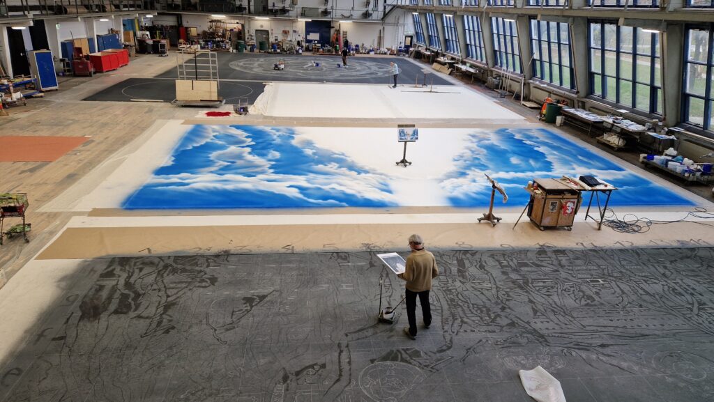 Eine große Werkhalle mit mehreren bemalten Bühnenhintergründen, darunter ein Gemälde von blauen Wolken, und eine Person bei der Arbeit.