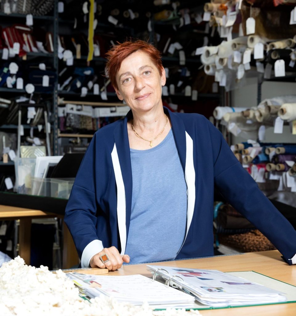 Eine Frau mit kurzen roten Haaren steht vor Tischen mit Stoffproben und einem offenen Ordner in einem Stofflager.