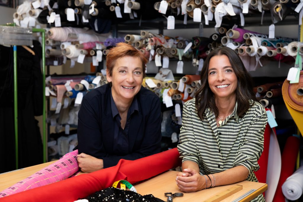Two smiling women sit at a table in a fabric workshop, surrounded by rolls of fabric and tailoring accessories.