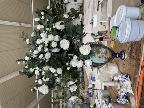 Große Vase mit gemaltem Motiv und weißen Blumen, umgeben von Malutensilien auf einem Holztisch in einem Atelier.