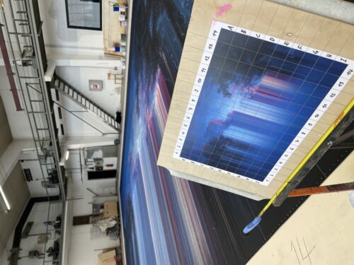 Ein großes Gemälde mit bunten Linien liegt in einer Atelierhalle, daneben ein Raster mit einer Miniaturversion des Motivs.