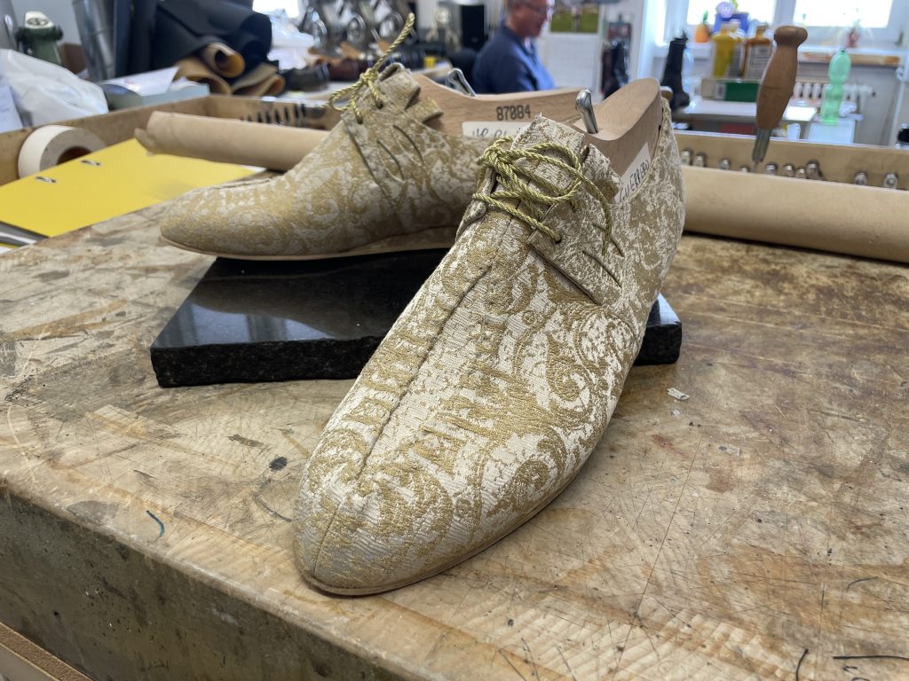 A pair of elegant, patterned lace-up shoes with gold and white decorations stands on a workbench in a workshop.