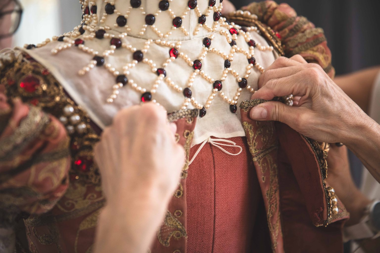 Hände helfen dabei, ein aufwendig verziertes historisches Kostüm mit Perlen und roten Steinen anzuziehen oder zu verschließen.