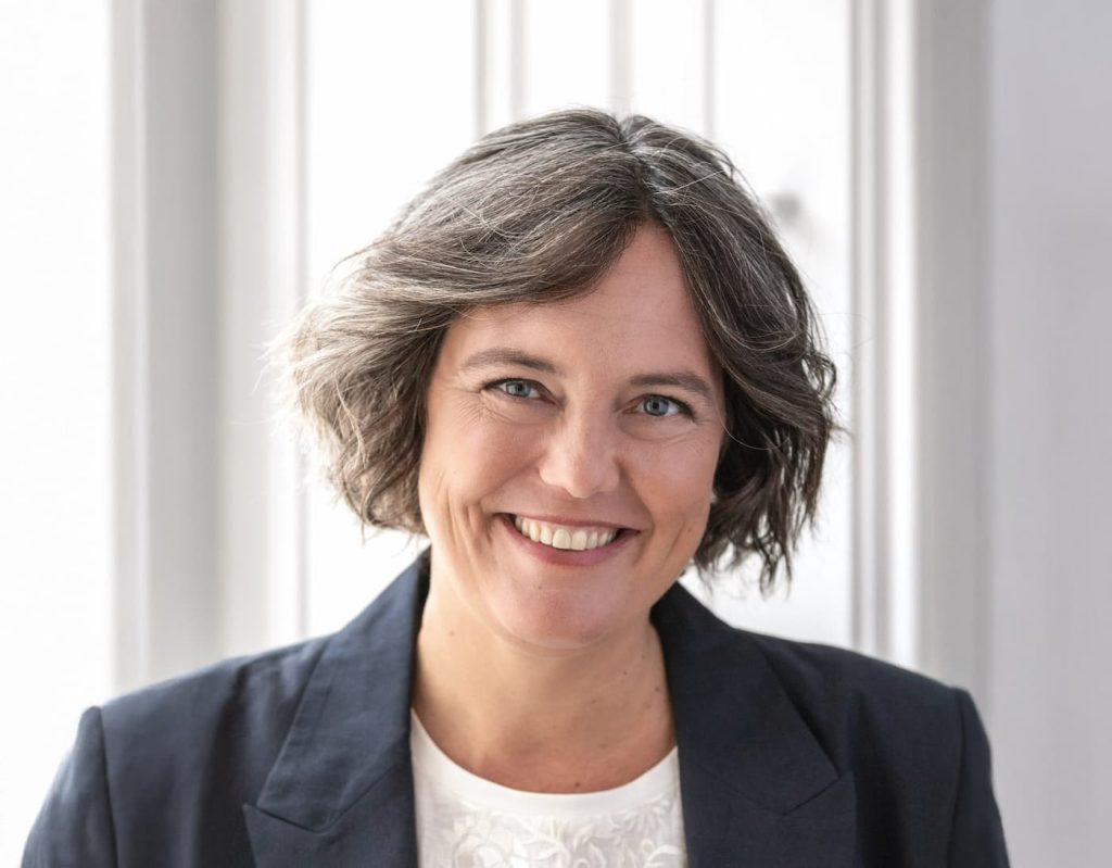 Frau mit schulterlangem, welligem, graubraunem Haar und dunkelblauem Blazer lächelt vor einem hellen Fenster.