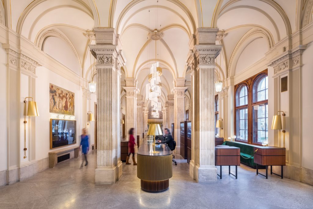 Großzügige Halle mit hohen, gewölbten Decken, verzierten Säulen und mehreren Personen in Bewegung, elegante Beleuchtung und Dekor.