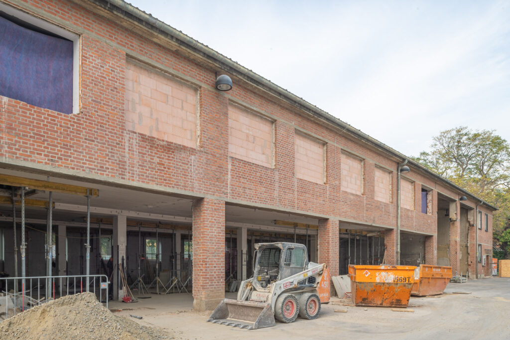 A brick building under reconstruction with open ground floor areas, an excavator and two construction containers in front of it.