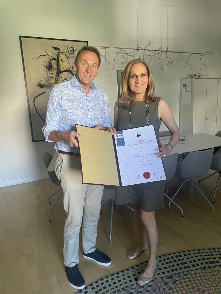 A woman and a man stand next to each other in an office, both holding a certificate together and smiling at the camera.