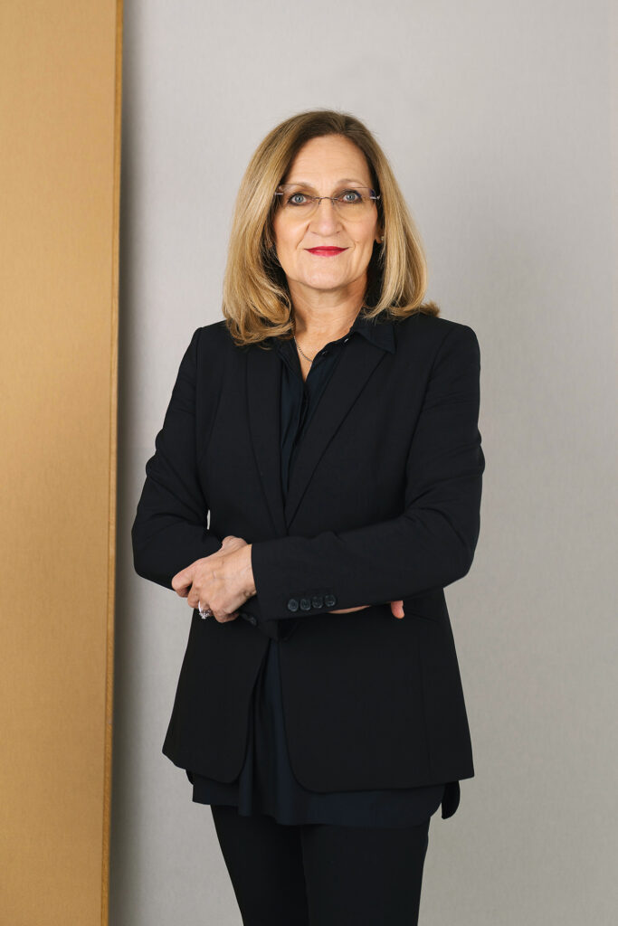 Frau mit schulterlangem blonden Haar, Brille und dunklem Blazer steht mit verschränkten Armen vor neutralem Hintergrund.