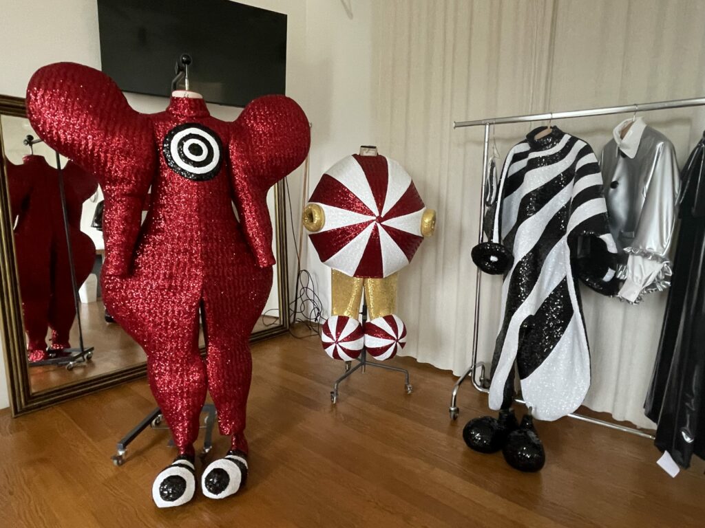 Three colourful, eye-catching costumes, one red with large circles, one with round stripes and one black and white with glitter, stand on clothes racks.