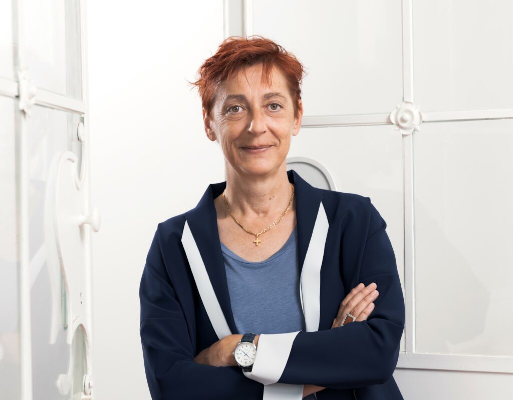 Frau mit kurzem, rötlichem Haar und verschränkten Armen, trägt ein dunkelblaues Sakko und steht vor einem weißen Hintergrund.