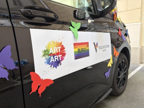 Ein schwarzes Auto mit bunten Papier-Schmetterlingen und einem Schild mit den Logos von ART for ART, Burgtheater und Volksoper Wien.