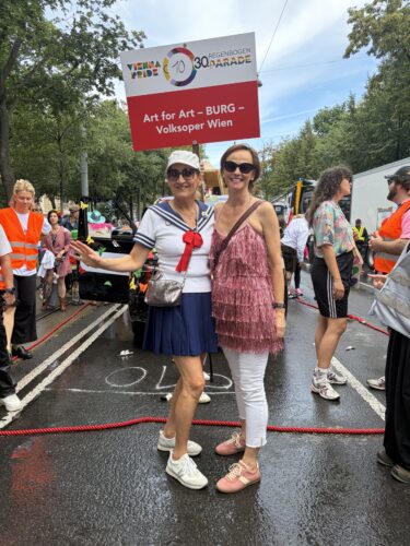 Zwei Frauen posieren lächelnd auf einer Straße, eine trägt ein Matrosenkostüm, die andere ein rosa Fransenkleid; Menschen und ein Schild im Hintergrund.