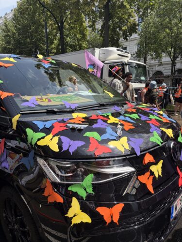 Schwarzes Auto mit bunten Papier-Schmetterlingen bedeckt, im Hintergrund Menschen und Bäume bei einer Parade oder Demonstration.