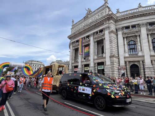 Ein Umzugswagen mit bunten Papierdreiecken und Regenbogenflaggen fährt bei einer Pride-Parade vor einem historischen Gebäude vorbei.