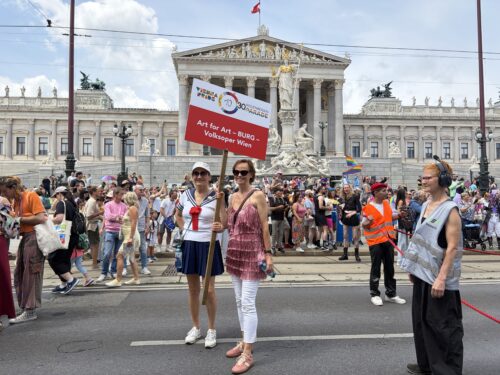 Zwei Frauen halten ein Schild bei einer Parade vor dem österreichischen Parlament, umgeben von einer bunten Menschenmenge.