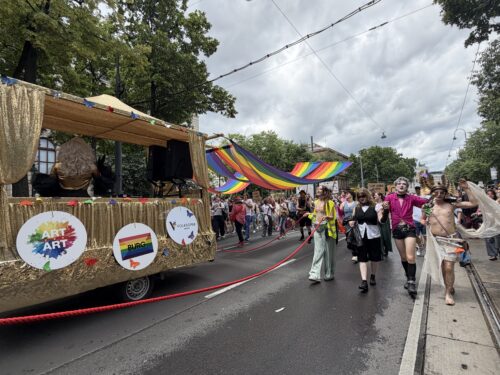 Menschen nehmen an einer bunten Parade mit Regenbogenflaggen und einem geschmückten Wagen bei bewölktem Himmel teil.