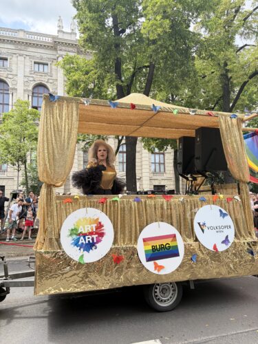 Eine Dragqueen steht auf einem gold geschmückten Wagen bei einer Parade, mit Schildern „Art“, „Burg“ und „Volksoper Wien“.