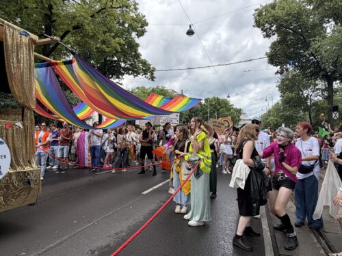 Menschen feiern auf einer Straße während einer Parade mit bunten Regenbogenflaggen und festlicher Dekoration.