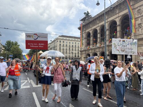 Menschen nehmen an einer bunten Parade mit Regenbogenfahnen und Transparenten vor einem historischen Gebäude in Wien teil.