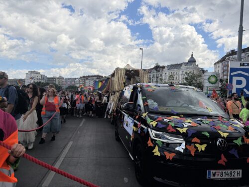 Ein mit bunten Schmetterlingen geschmücktes Auto fährt vor einer Menschenmenge bei einer bunten Parade auf einer Straße.