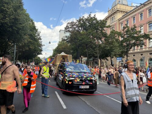 Menschen marschieren bei einer Parade auf einer Stadtstraße, begleitet von einem geschmückten Auto und bunt gekleideten Teilnehmern.