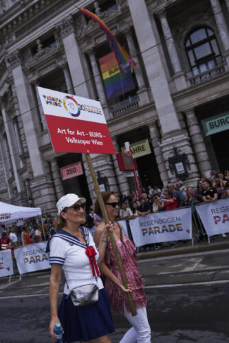 Zwei Frauen gehen bei einer Regenbogenparade, eine trägt ein Schild der Volksoper Wien, im Hintergrund Regenbogenflaggen und Zuschauer.
