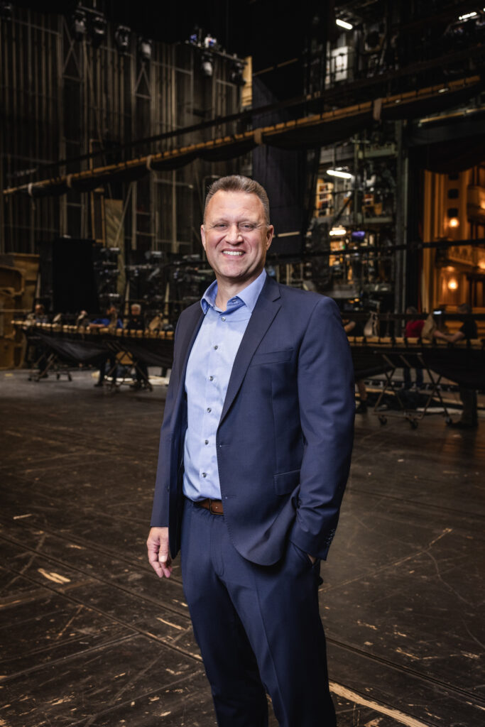 Ein Mann im Anzug steht lächelnd auf einer Theaterbühne mit Technik und Kulissen im Hintergrund.